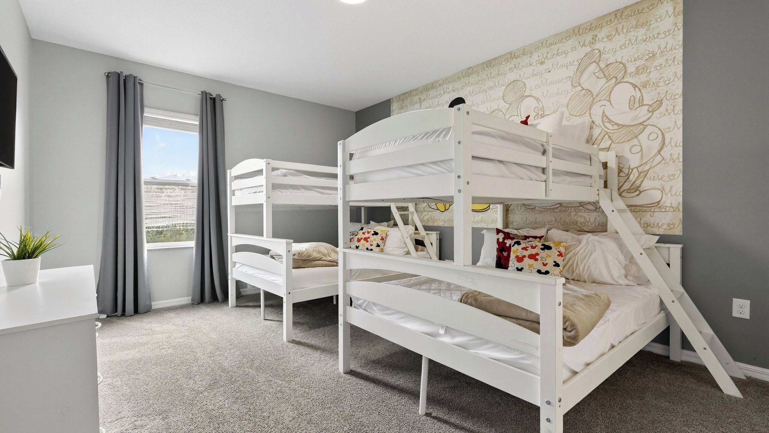 A room with bunk beds, a window with curtains, a wall with a Mickey Mouse mural, and a small plant on a table.