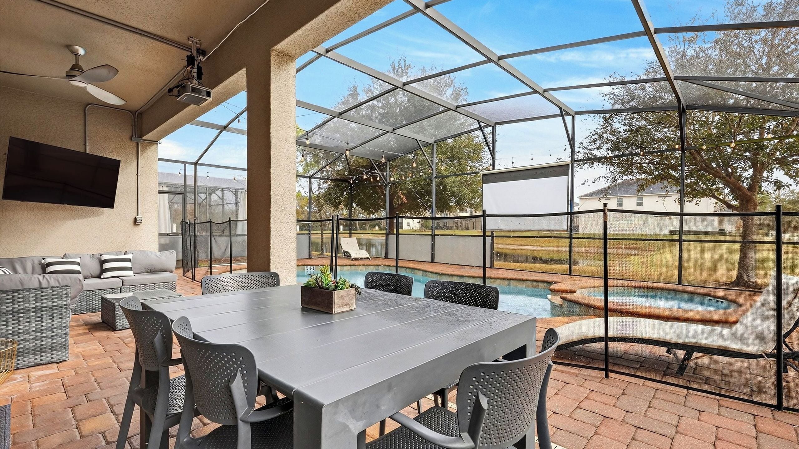 A covered patio with a table and chairs, a TV mounted on the wall, and a view of a swimming pool.
