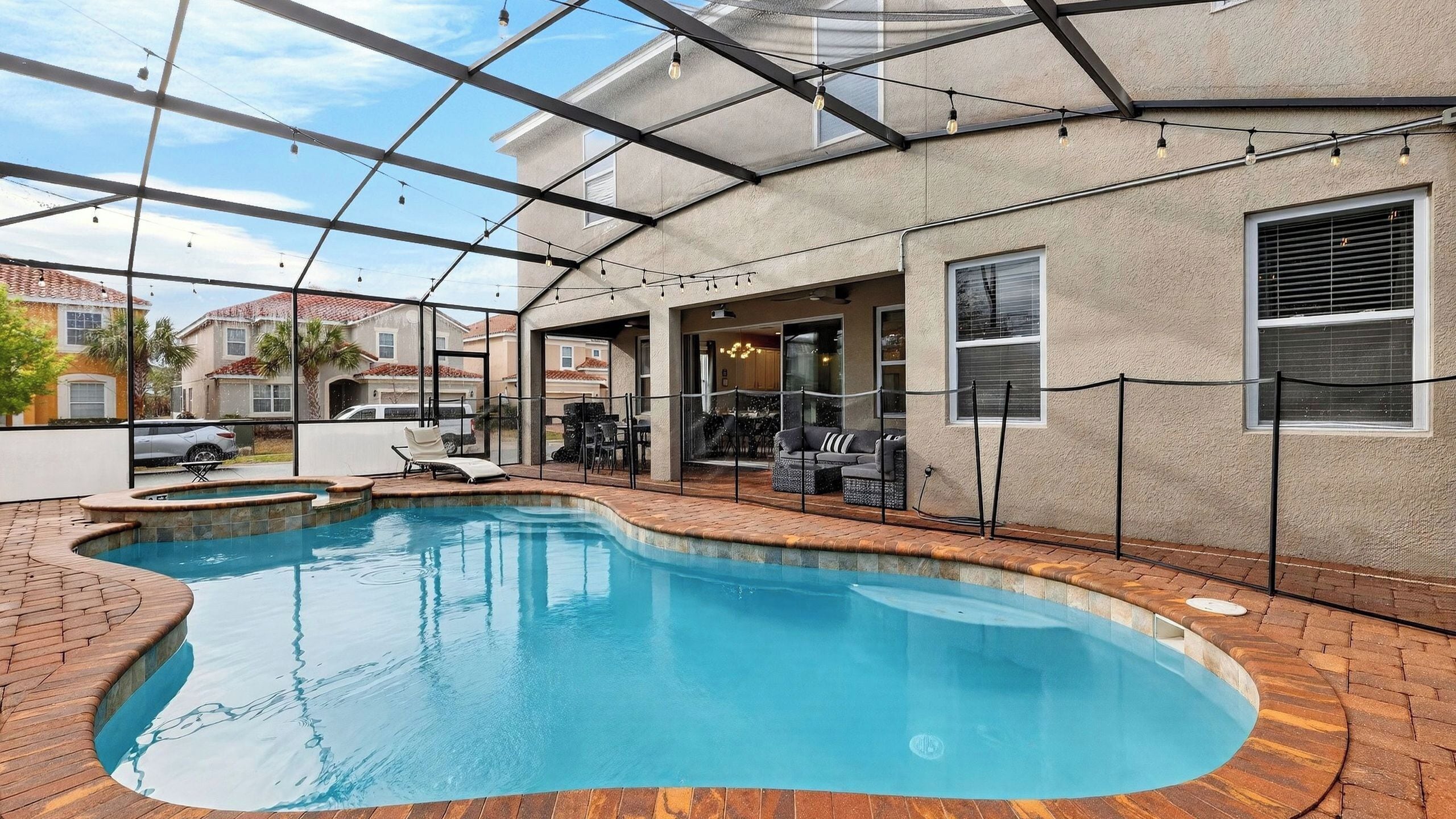 A swimming pool with a brick deck, a covered patio area, and a view of neighboring houses.