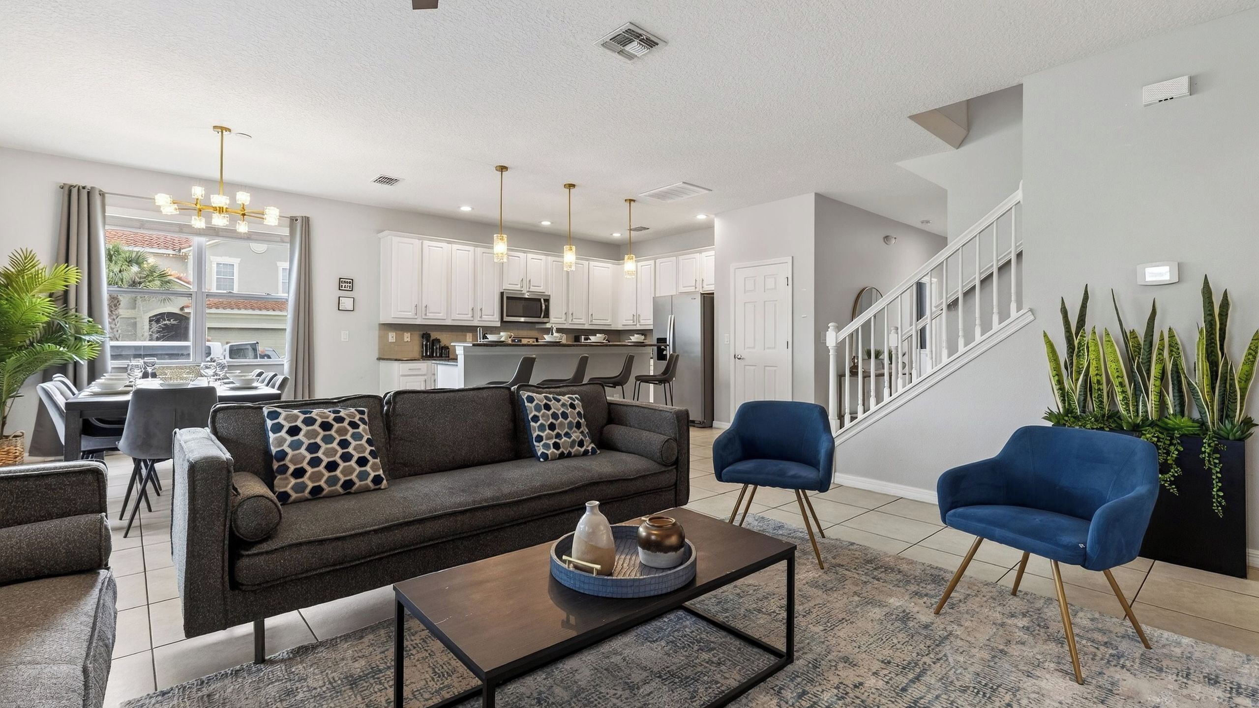 A modern living room with a sofa, coffee table, and blue chairs. A kitchen is visible in the background with white cabinets and stainless steel appliances.