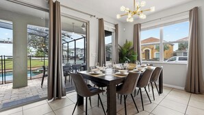 A dining area with a large table set for six, surrounded by chairs, with a view of a swimming pool and houses outside. - Shiva by Avantstay Sleeps 22 Pool + Hot Tub + Great Park Access (Kissimmee)