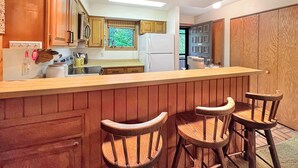 Une cuisine avec des meubles en bois, un îlot central doté de tabourets, un réfrigérateur et une fenêtre avec des stores.