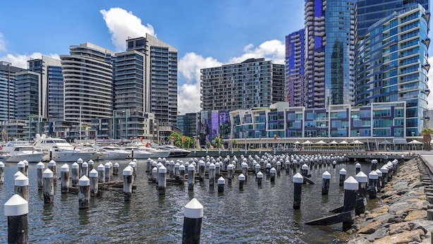 A waterfront cityscape with modern high-rise buildings, a marina with yachts, and a dock with bollards. - KozyGuru | Docklands | Waterfront Skyline Retreat (Docklands)