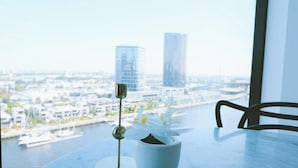 A table with a potted plant and a glass of water, overlooking a cityscape with buildings and a river. - KozyGuru | Docklands | Waterfront Skyline Retreat (Docklands)