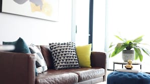 A brown leather sofa with patterned cushions, a blue ottoman, and a potted plant on a small table. - KozyGuru | Docklands | Waterfront Skyline Retreat (Docklands)