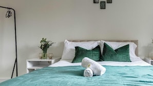 A neatly made bed with a teal bedspread, white pillows, and a rolled towel. There are bedside tables with small plants and a wall-mounted picture frame. - Stylish 1-Bed Apartment in Croydon | Easy Access to Central London (South Croydon)
