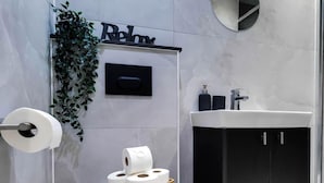 A modern bathroom with a toilet, a sink, a black cabinet, a wall-mounted toilet paper dispenser, and a decorative plant. - Stylish 1-Bed Apartment in Croydon | Easy Access to Central London (South Croydon)