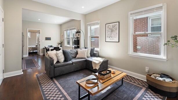 A living room with a gray sectional sofa, a wooden coffee table, and a large window with a view of a brick building. - Beltline Cottage • Walk to Ponce City Market (Atlanta)