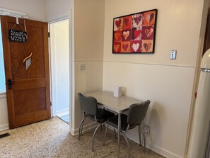 Un coin repas avec une table et des chaises, une porte en bois avec un meuble à chaussures, et une œuvre d’art sur le thème du cœur accrochée au mur.