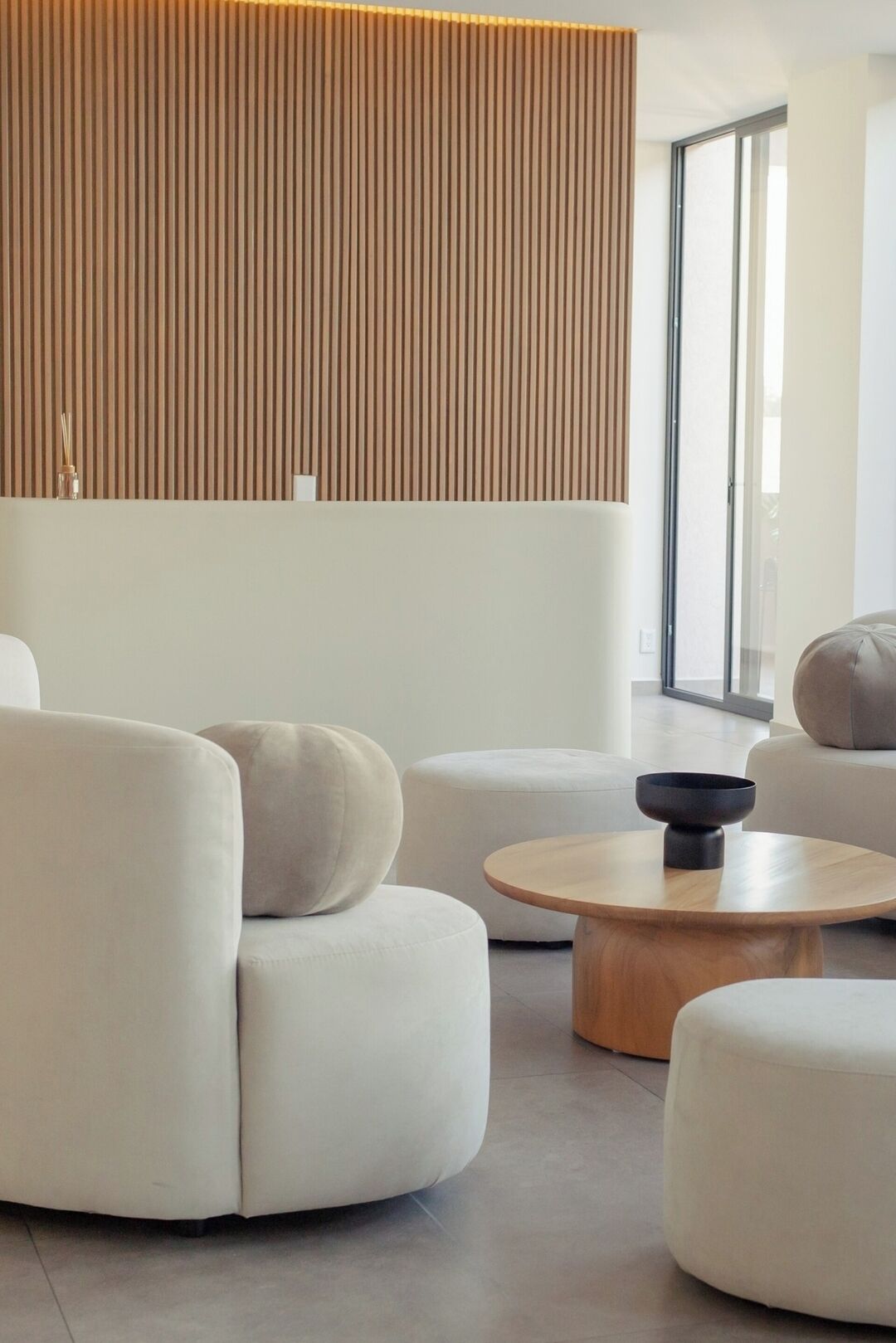 A modern living room with a sofa, ottoman, and coffee table, a wooden slatted wall, and a large window.