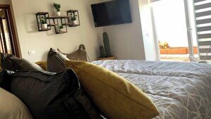 Ein Schlafzimmer mit Bett, einem an der Wand montierten Fernseher, einem Kaktus und einem Balkon mit Pflanzen.