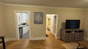 A living room with hardwood flooring, a television on a wooden stand, and a kitchen visible through an open door.