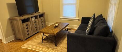 A living room with a wooden coffee table, a dark sofa, a TV on a wooden cabinet, and a window with blinds.