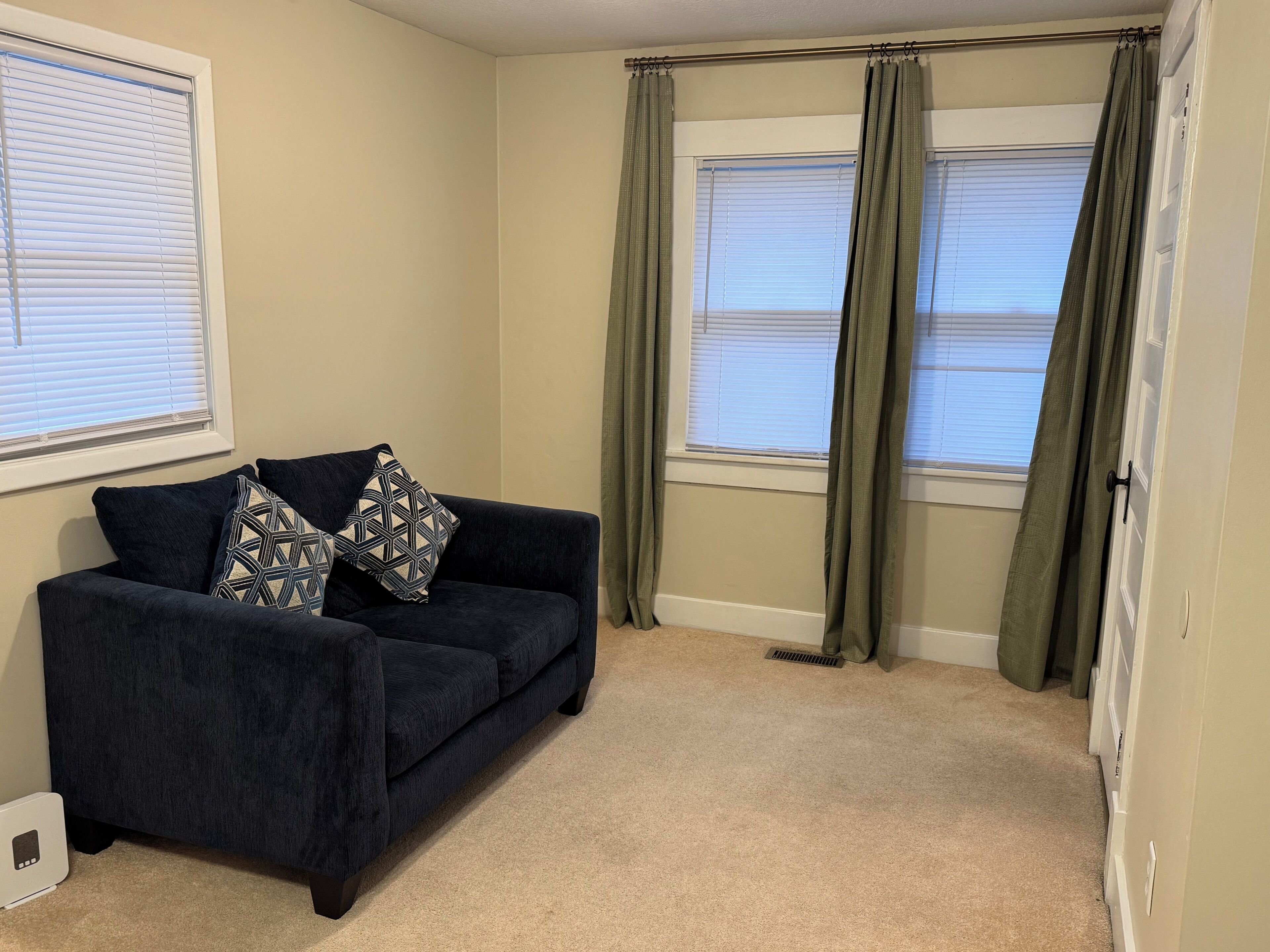 A room with a dark blue sofa, patterned cushions, beige carpet, and a window with green curtains.