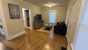 A living room with hardwood flooring, a TV on a stand, a sofa, and a dining table.