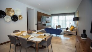 Ein modernes Wohnzimmer mit einem blauen Sofa, einem Essbereich mit Tisch für vier Personen und einer Küchenzeile mit Holzschrank und großem Fenster.