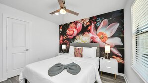 A bedroom with a large floral mural, a bed with a bow-shaped pillow, a ceiling fan, bedside lamps, and a white nightstand. - Arbor Flats #8 in the Heart of Houston (Houston)