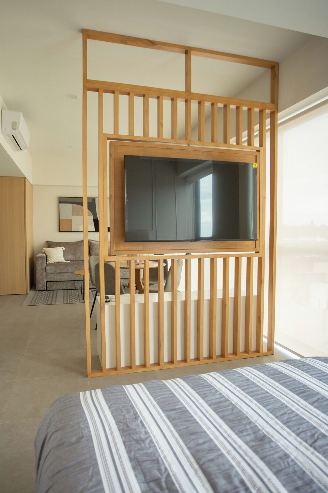 A bedroom with a bed, a TV mounted on a wooden partition, a sofa, and a window.