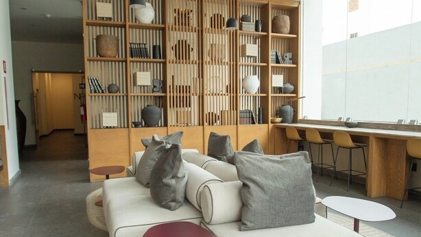 Uma sala de estar moderna com um sofá seccional, uma mesa lateral vermelha, uma estante de madeira com diversos objetos decorativos e iluminação pendente.