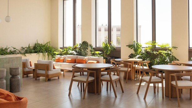 Un espace intérieur vaste et lumineux, avec plusieurs tables et chaises rondes, de grandes fenêtres et des plantes d’intérieur.