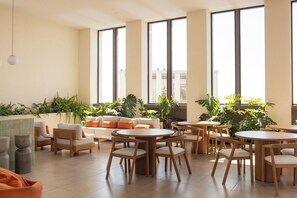 Un espacio interior amplio con varias mesas redondas y sillas, ventanas grandes y plantas de interior.