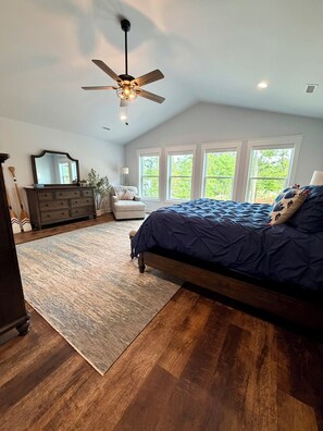 A bedroom with a large bed, a ceiling fan, a dresser, a chair, and a view of trees through the windows. - Cozy Ridge Retreat: A Spacious 6-bed waterfront house on beautiful Lake Mitchell (Rockford)