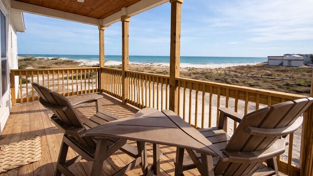 Terraza de madera con una mesa y sillas, con vista a una playa con dunas y cielo despejado.