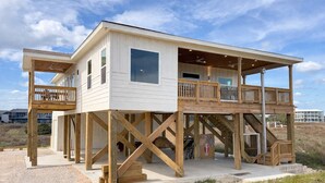 Una casa de playa de dos pisos con balcón, terraza de madera y una escalera.