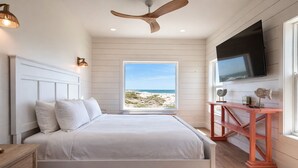 Un dormitorio con cama, ventilador de techo, televisor, escritorio de madera y ventana con vista al mar.