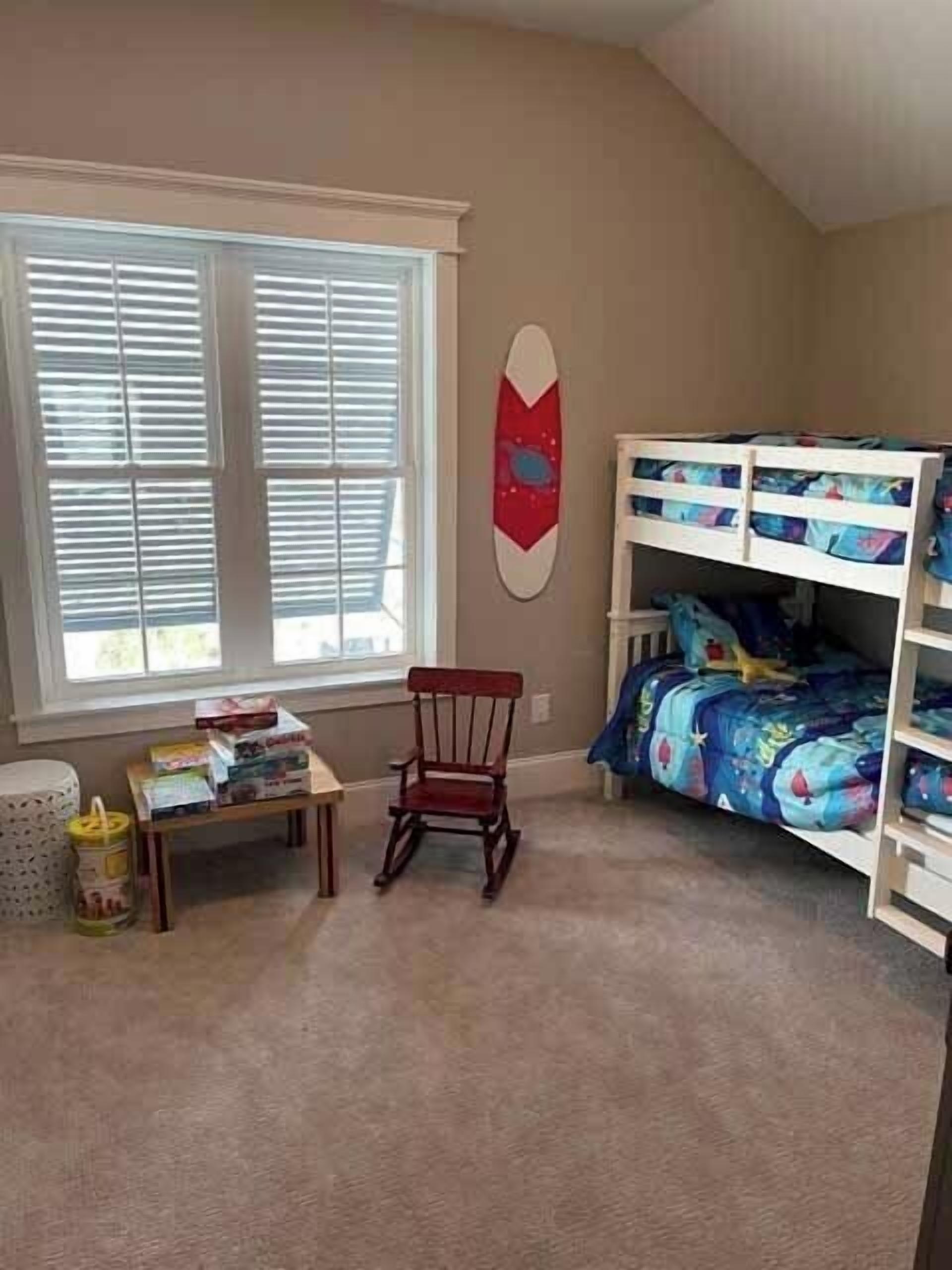 A child's bedroom with a bunk bed, a rocking chair, a small table with toys, and a window with blinds.