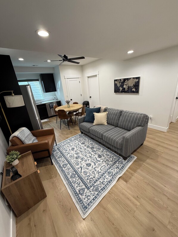 A modern living room with a sofa, a coffee table, a rug, and a dining area with a table and chairs. - 2-bedroom apartment in Utah County (American Fork)