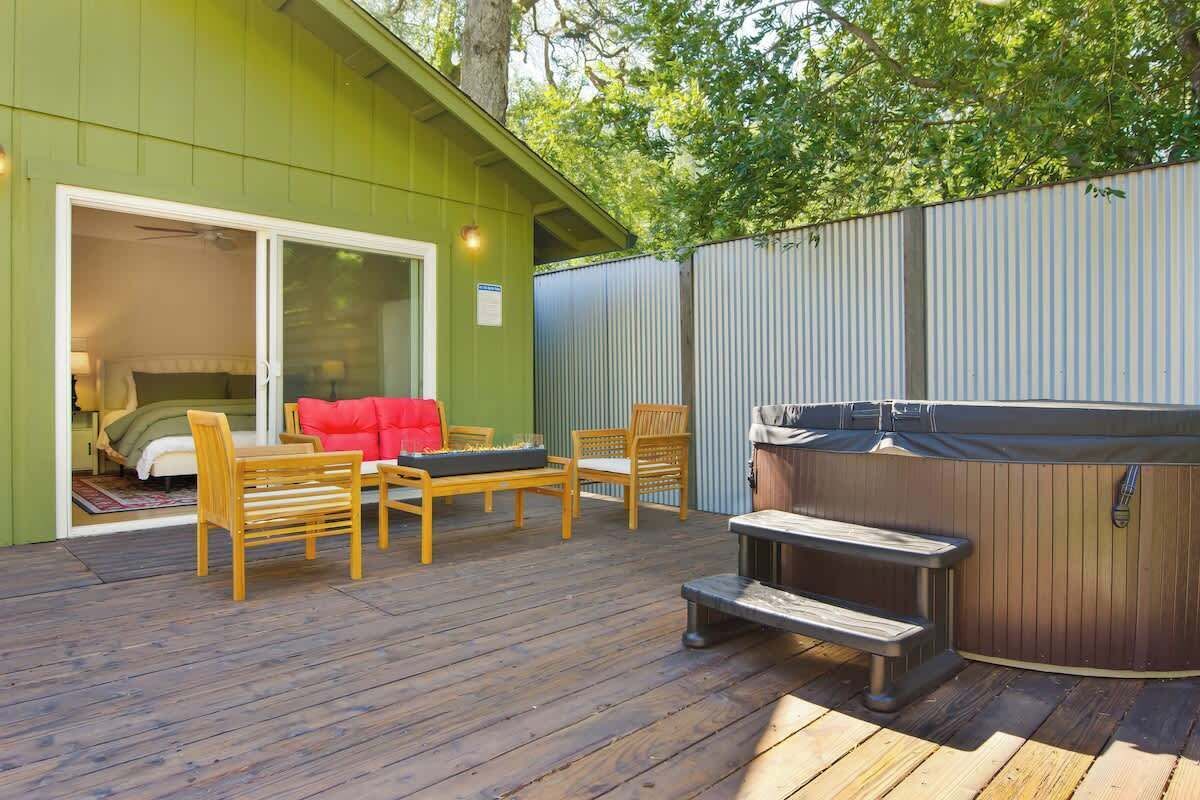 A deck with a hot tub, wooden furniture, and a sliding glass door leading to a bedroom.