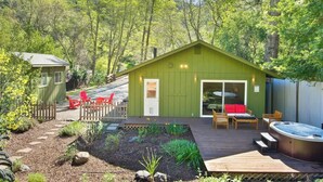 A green cabin with a hot tub, deck, and outdoor seating area surrounded by a garden and forest. - Wine Country Creekside Cottage Bridge & Day Studio (Kenwood)