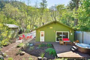 A green cabin with a hot tub, deck, and outdoor seating area surrounded by a garden and forest. - Wine Country Creekside Cottage Bridge & Day Studio (Kenwood)