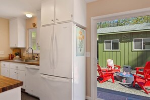 A kitchen with white cabinets, a refrigerator, and a dining area with red chairs and a fire pit. - Wine Country Creekside Cottage Bridge & Day Studio (Kenwood)