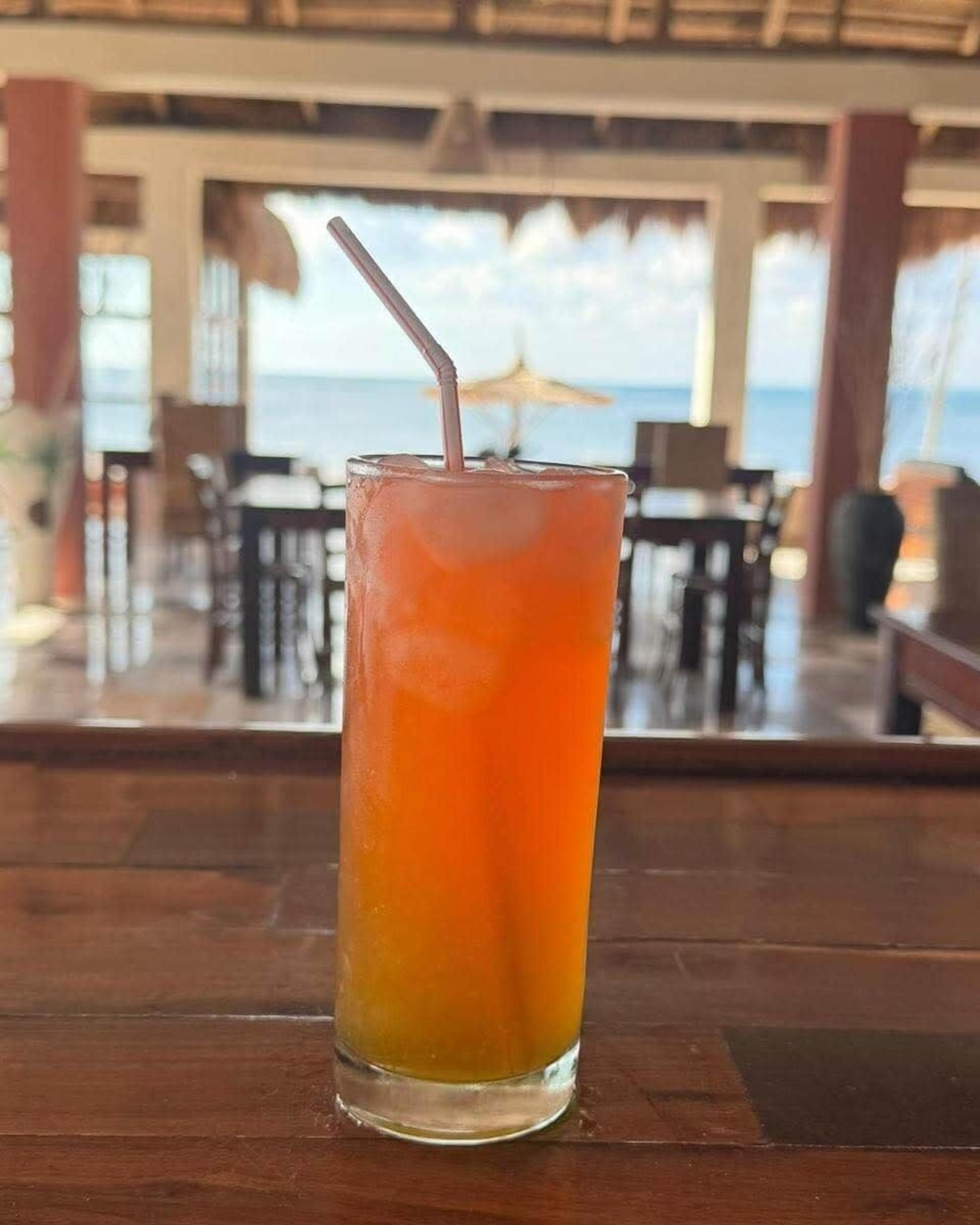 A tall glass with a straw, placed on a wooden surface, likely a bar or table, with a view of an outdoor seating area and a building in the background.