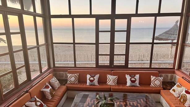 A room with a large window overlooking a beach, a sofa set, and a coffee table.