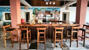 A bar area with wooden stools, a counter with various bottles, and a mural with a coconut theme.