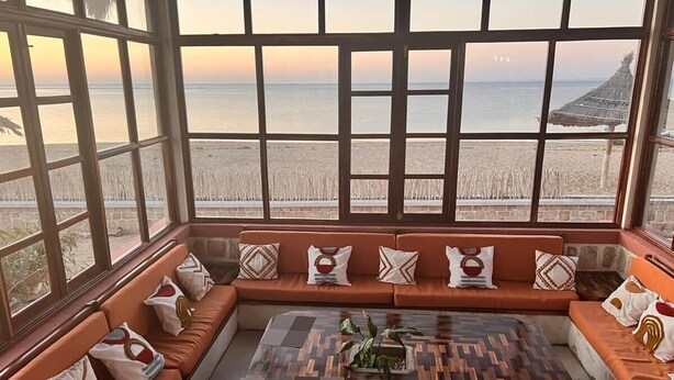 A room with a large window overlooking a beach, a wooden coffee table, and orange upholstered seating.