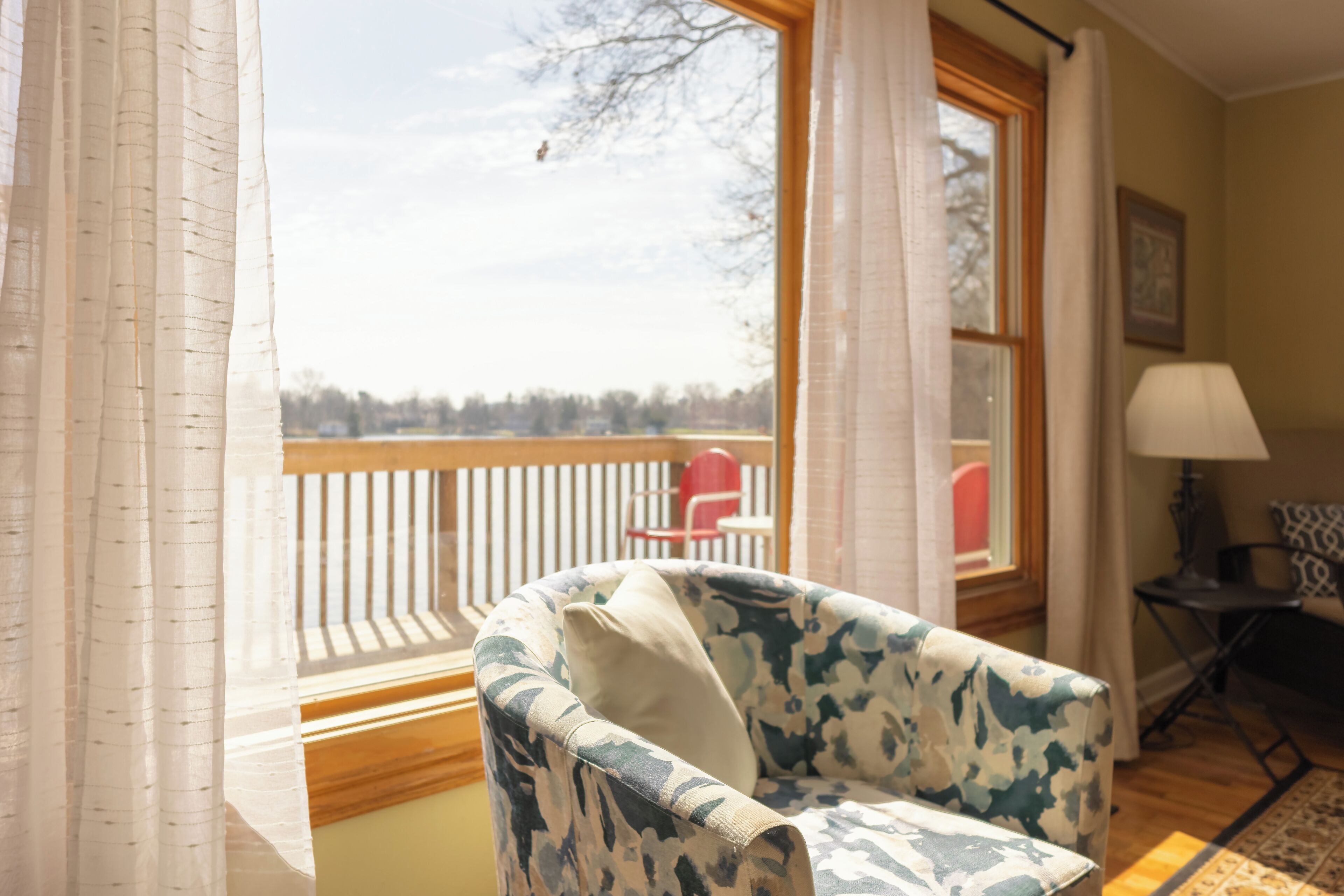 A room with a view through a window, a chair, a table, and a lamp.