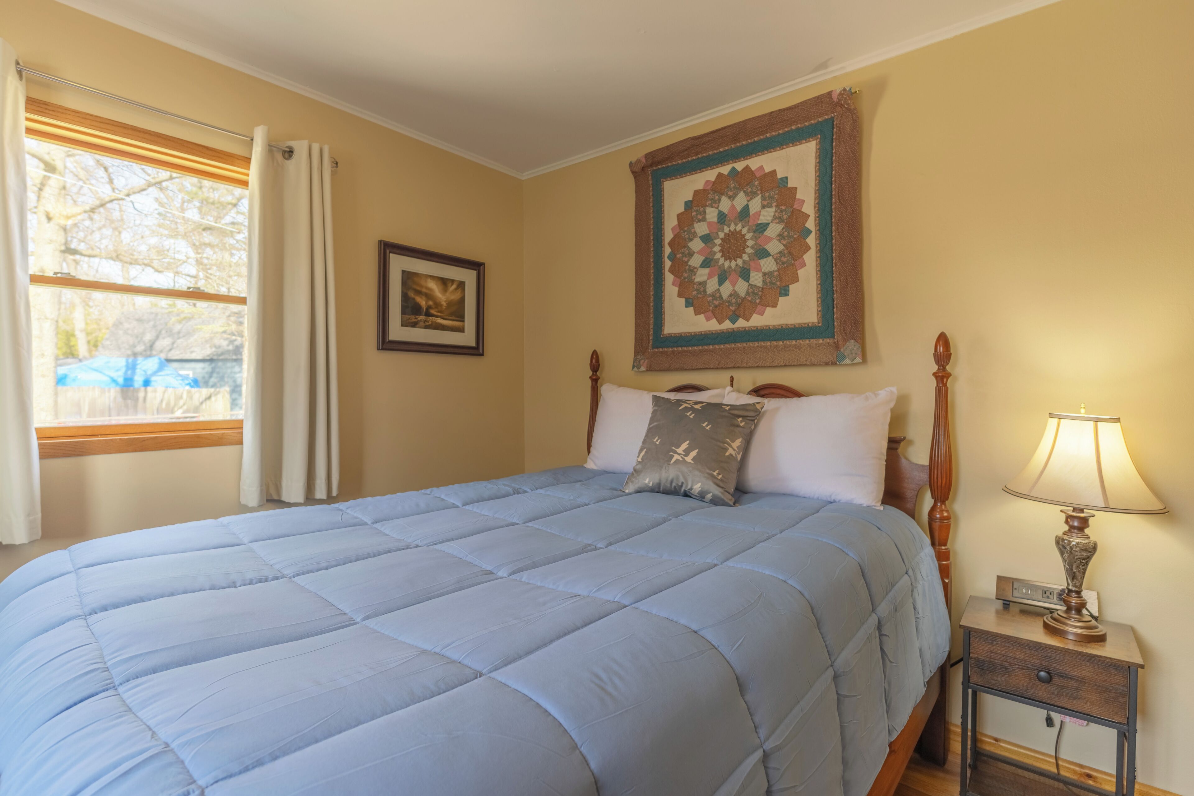 Un dormitorio con una cama grande, una mesita de noche con lámpara, una ventana con cortinas y un cuadro en la pared.