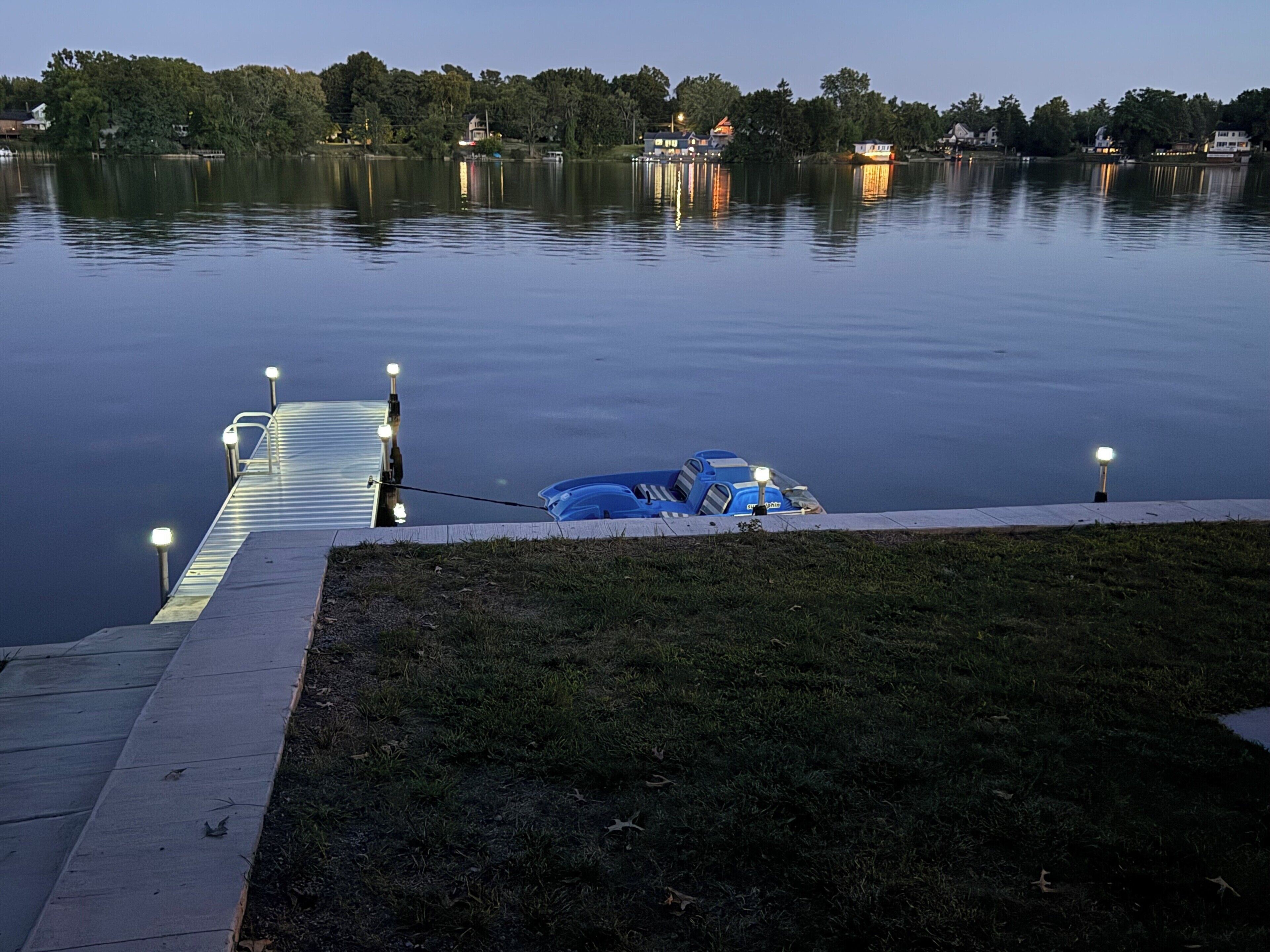 Ein ruhiger See mit einem Steg, der ins Wasser ragt, ein blaues Auto auf der Wiese geparkt und eine Reihe von Straßenlaternen entlang des Stegs.