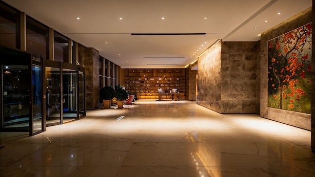 A modern lobby with a glass elevator, a seating area with tables, and a large wall with a painting. - Muwang Hotel Dalian (Dalian)