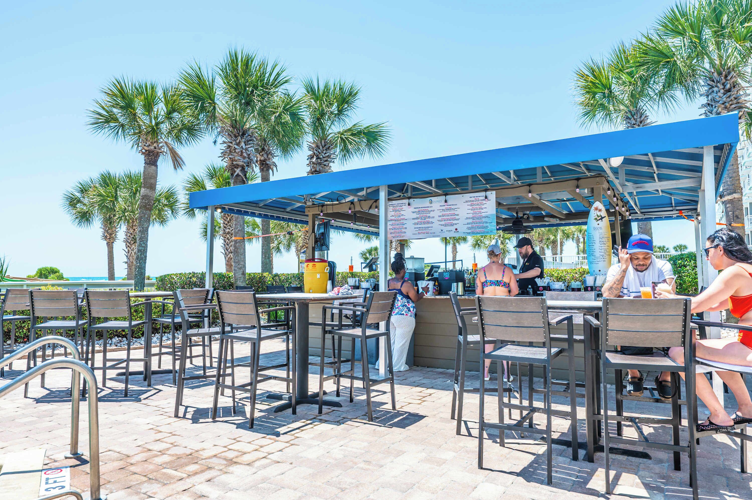 An outdoor seating area with tables and chairs, a covered section with a menu board, and palm trees in the background.