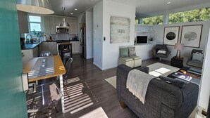 A modern kitchen with stainless steel appliances, a dining area with a wooden table and chairs, a grey sofa, and artwork on the walls.