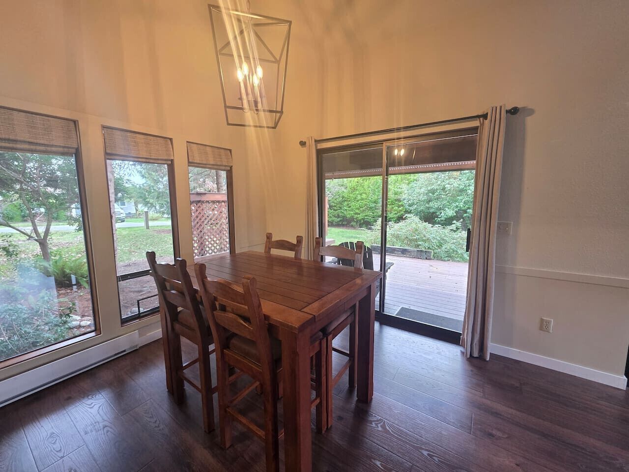 A dining area with a wooden table and chairs, large windows, and a view of greenery outside.