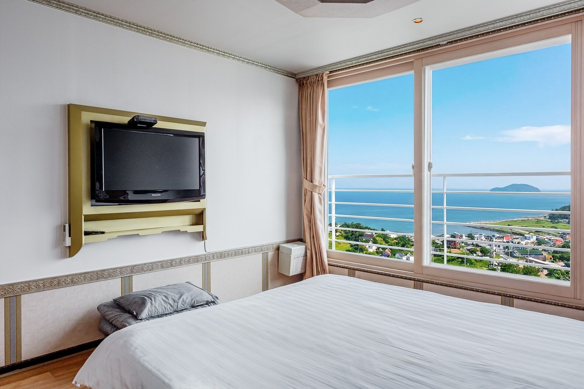 A hotel room with a bed, a mounted TV, a window with a view of the sea and a bridge, and a curtain.