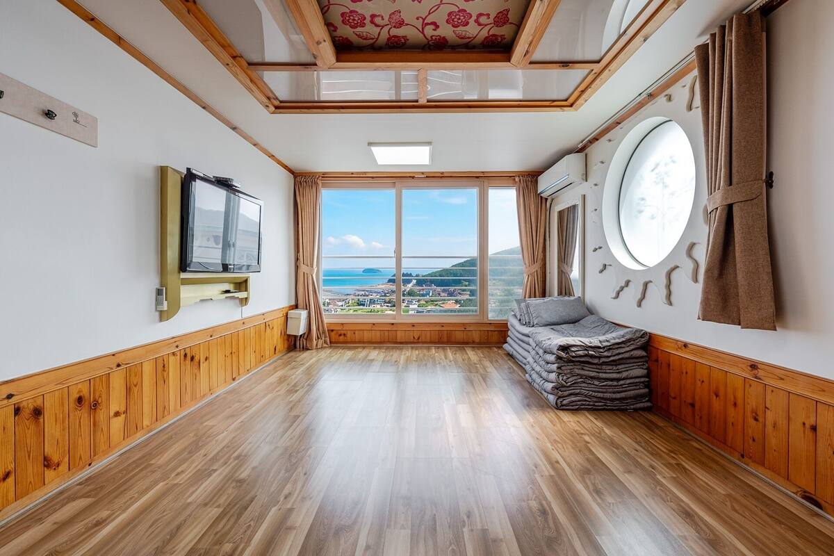 A room with wooden flooring, a television, a large window with a view, and a circular wall decoration.
