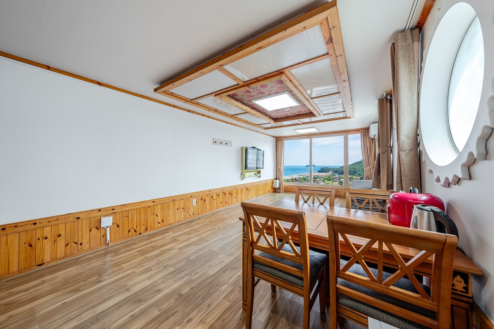 A room with a wooden floor, a dining table with chairs, a TV mounted on the wall, and a large window offering a view of the sea.