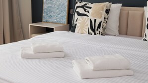 Un lit bien fait, avec deux serviettes blanches pliées, une tête de lit agrémentée de coussins décoratifs et une table de chevet sur laquelle est posée une œuvre d’art encadrée.